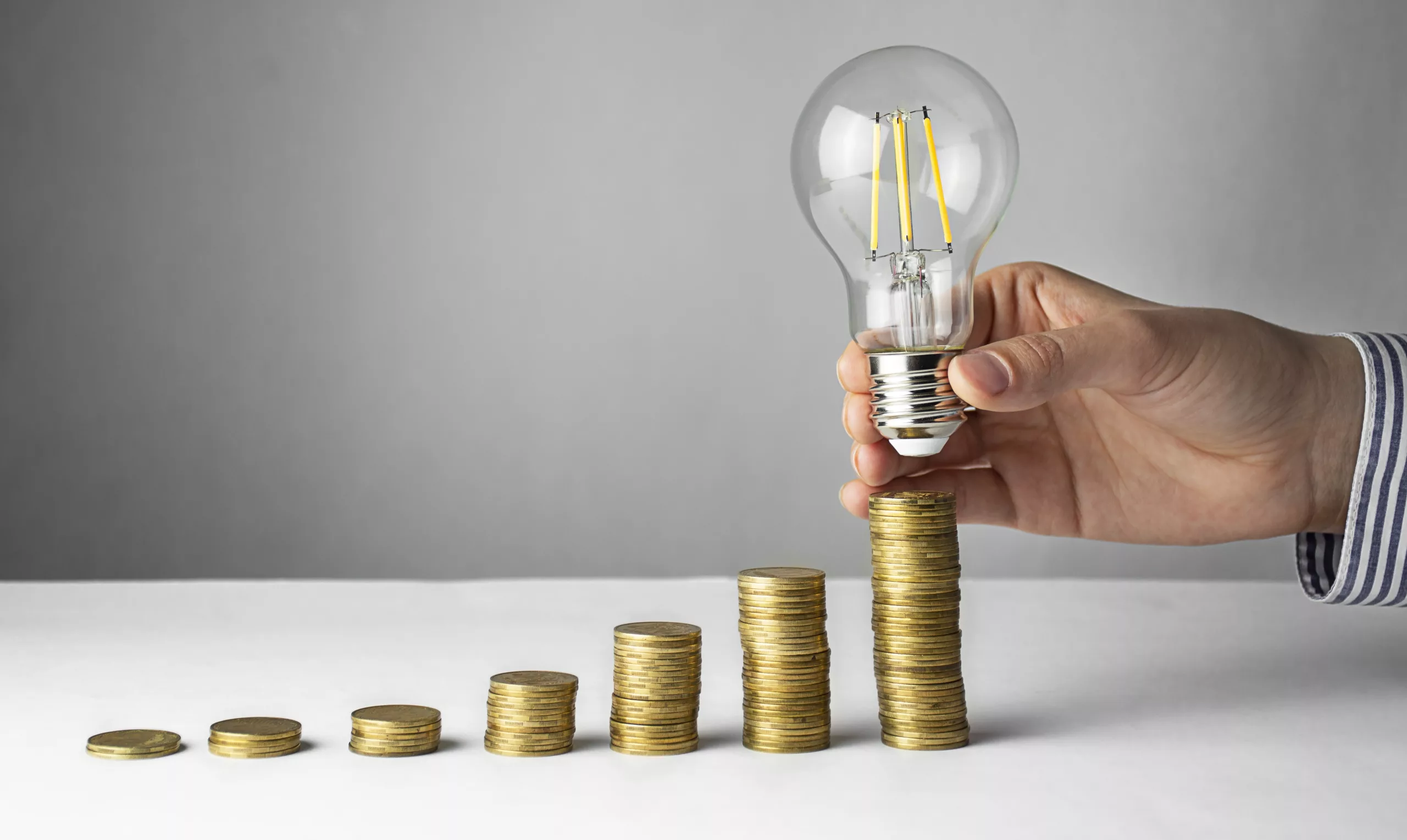 Business Concept. A Hand Holds A Lamp Over Coins On A Light Background. Business Ideas, Brainstorming. Recovery And Business Growth. Copy Space