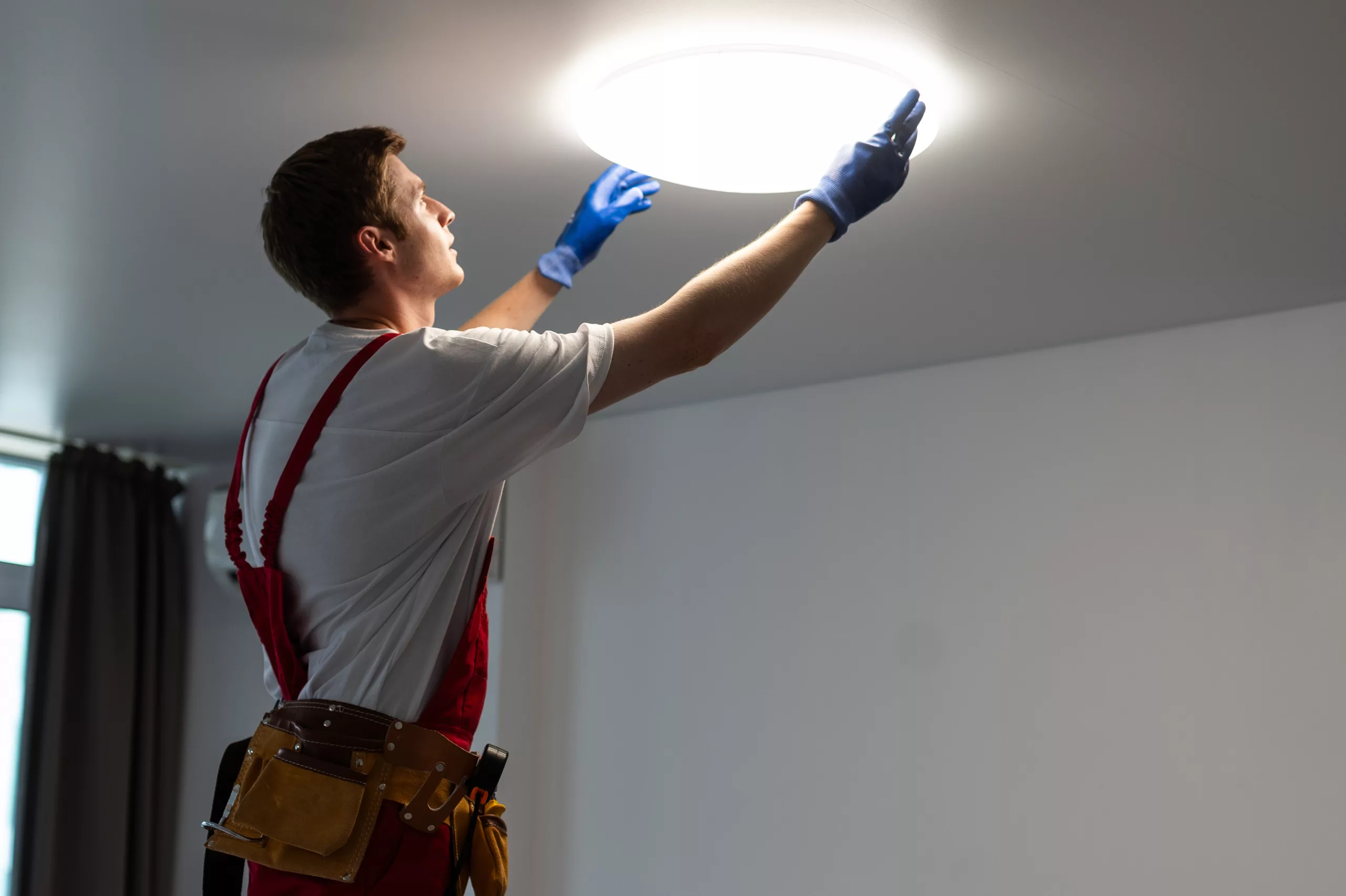 Electrician In Uniform Installing Ceiling Lamp Indoors. Space For Text