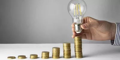 Business Concept. A Hand Holds A Lamp Over Coins On A Light Background. Business Ideas, Brainstorming. Recovery And Business Growth. Copy Space